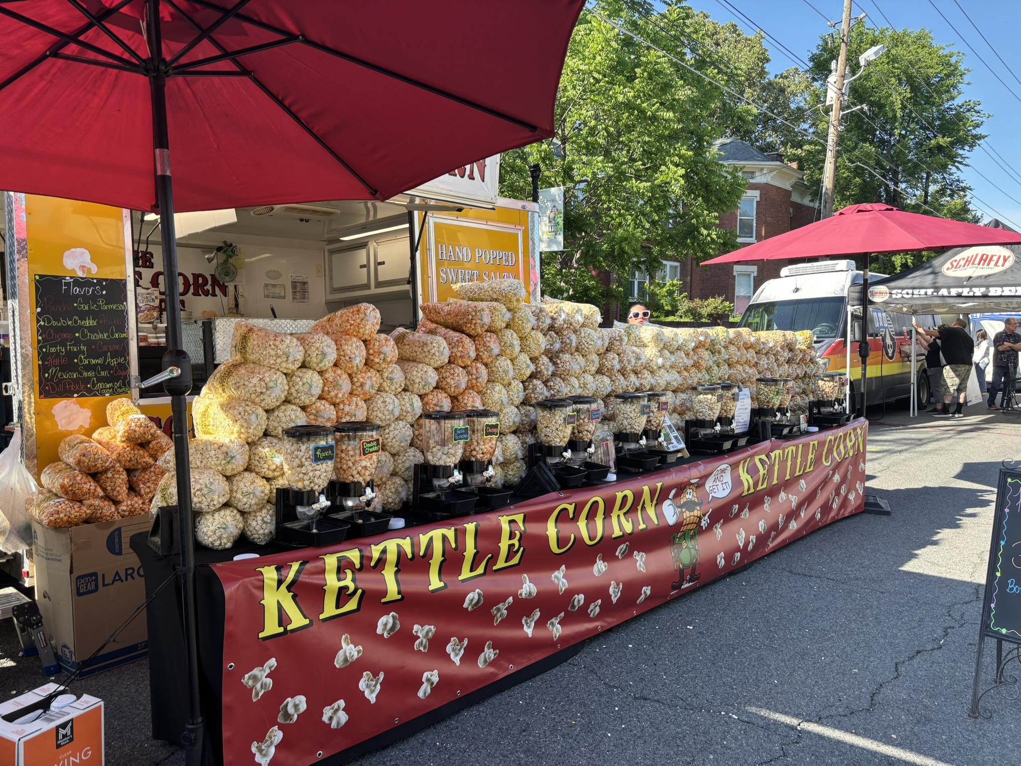 A long Kettle Corn Poppers table full of fresh bagged kettle corn and flavor containers.